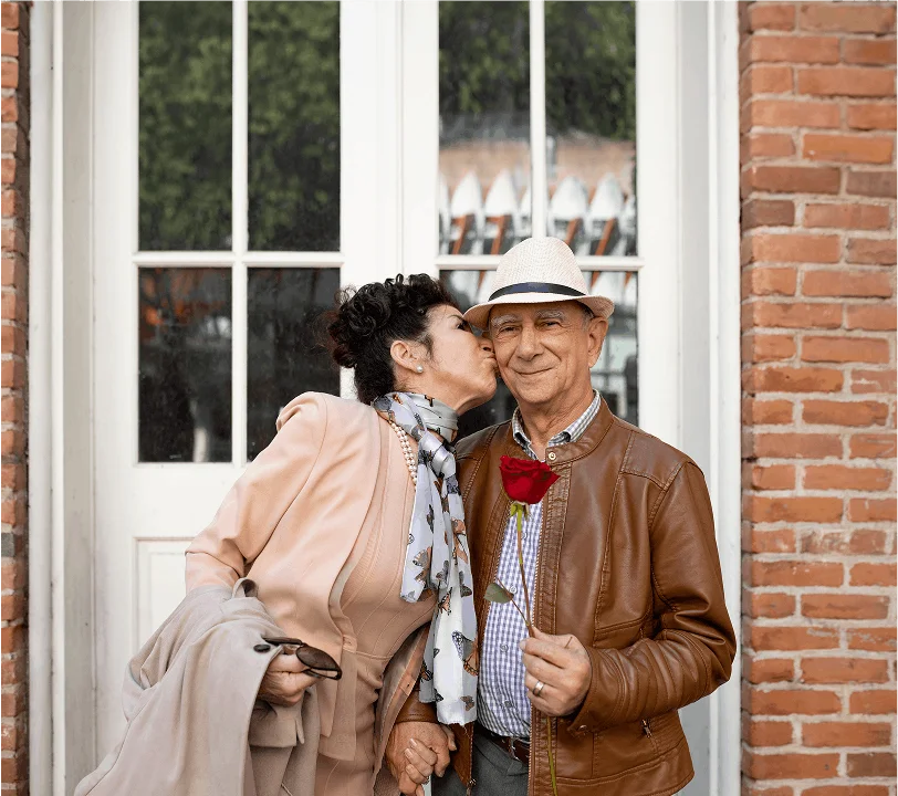 Older couple posing together outdoors, woman kissing man on the cheek while he holds a red rose